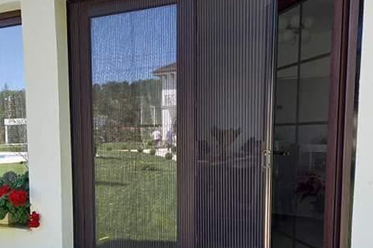 Plasa plisse maro, trasa pe jumatate peste o usa de terasa deschisa, cu pliurile panzei si manerul usii in prim-plan